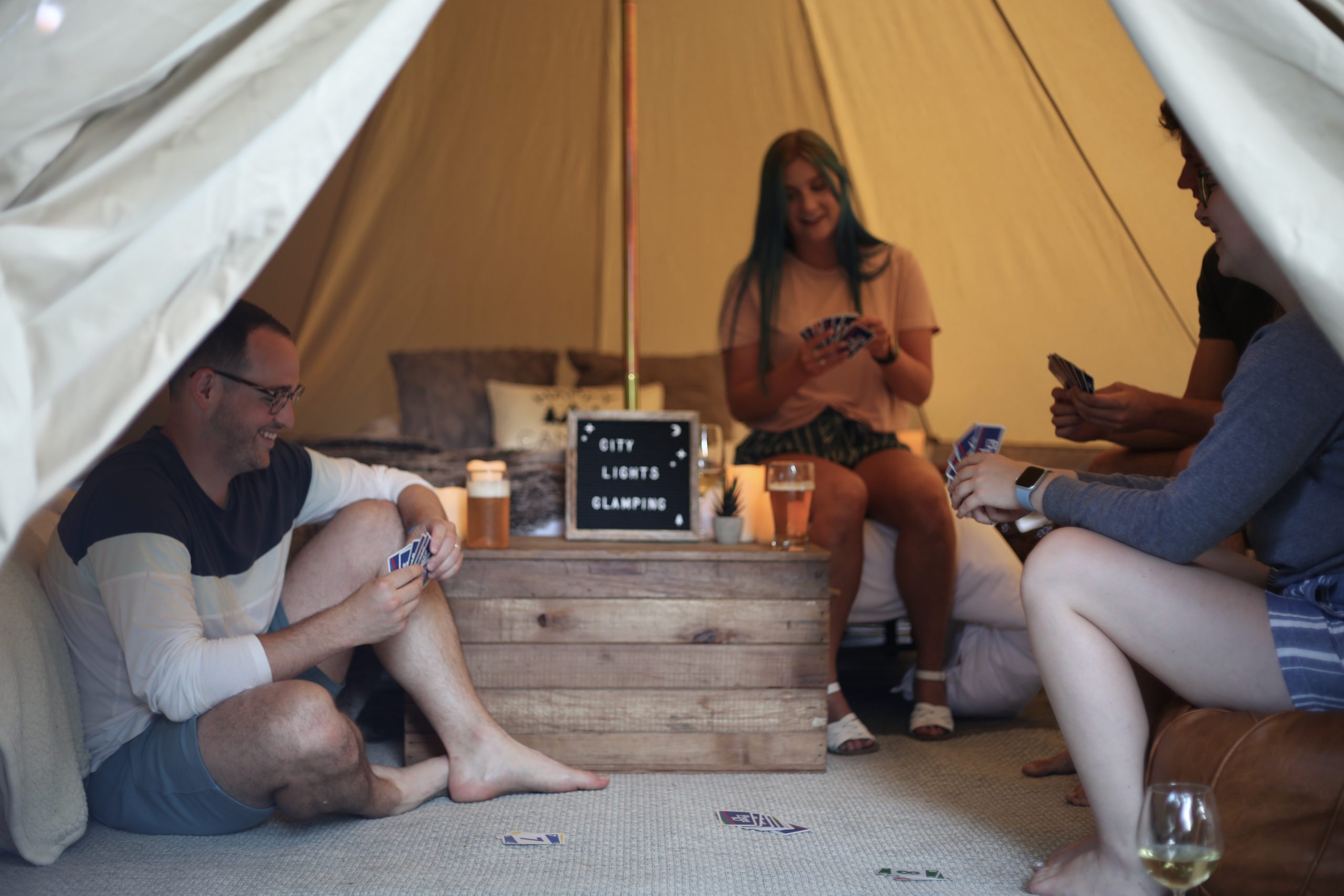 Four people sit inside a tent, playing cards around a wooden table with candles and a sign that reads "City Lights Glamping.