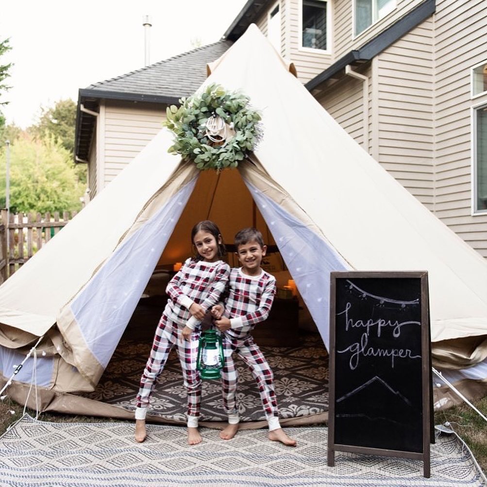 Two children in matching pajamas stand in front of a decorated outdoor tent, holding a lantern. A chalkboard sign nearby reads "happy glamping.
