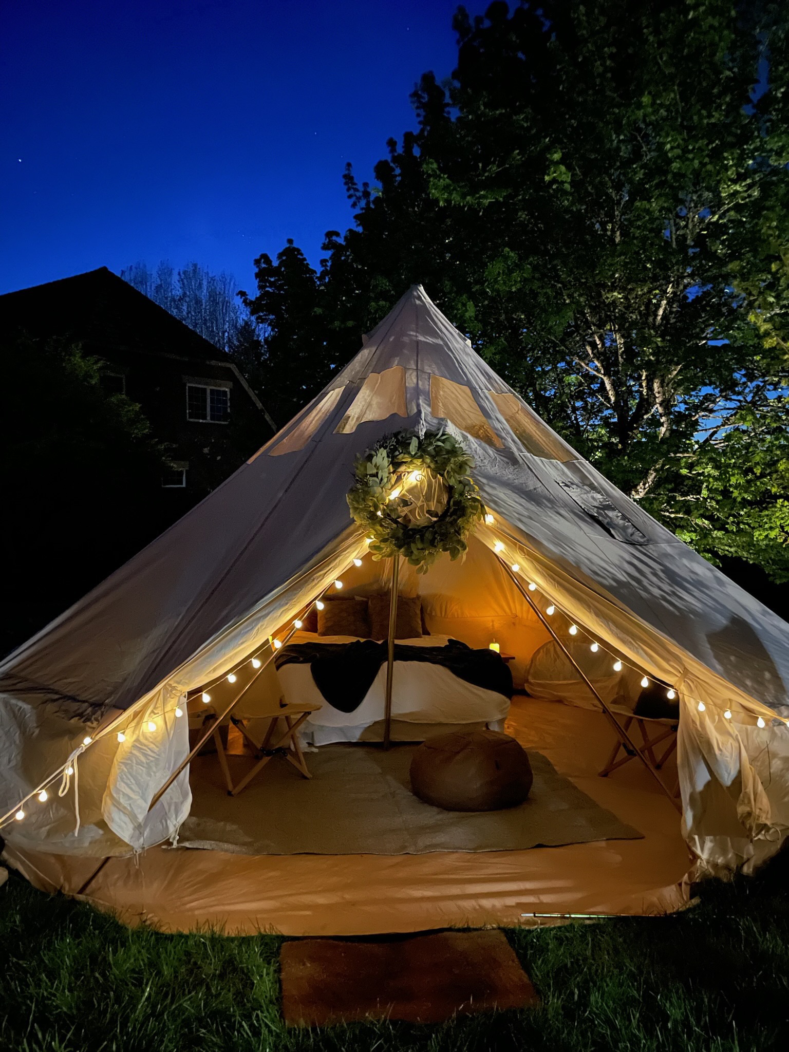 A canvas tent decorated with string lights and a wreath is set up at night, with a cozy interior visible, including a bed, table, and pouf.