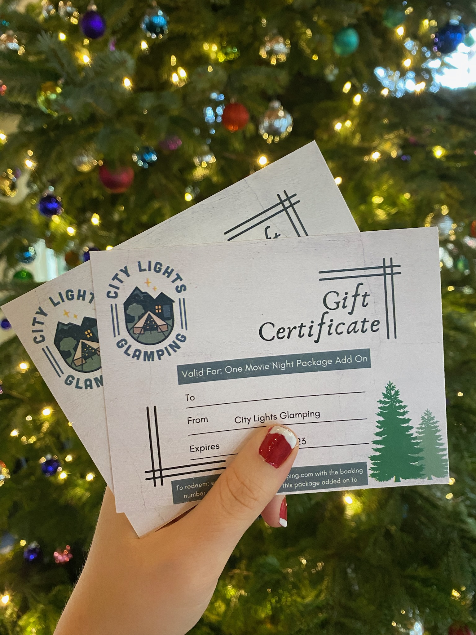 A hand holds two City Lights Glamping gift certificates in front of a decorated Christmas tree with lights and ornaments.