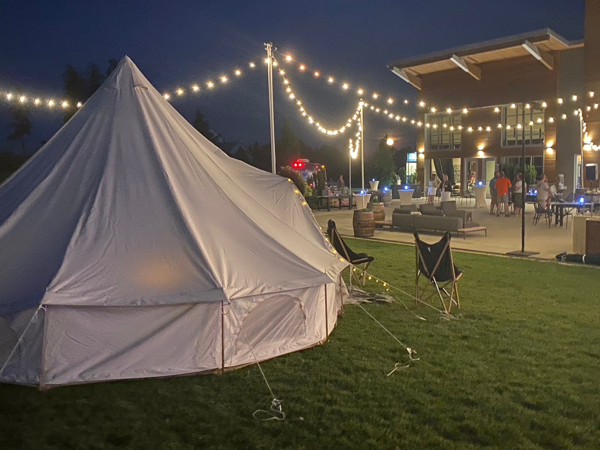 A large white tent and folding chairs are set up on grass near a building, with string lights overhead and people gathered outside at night.